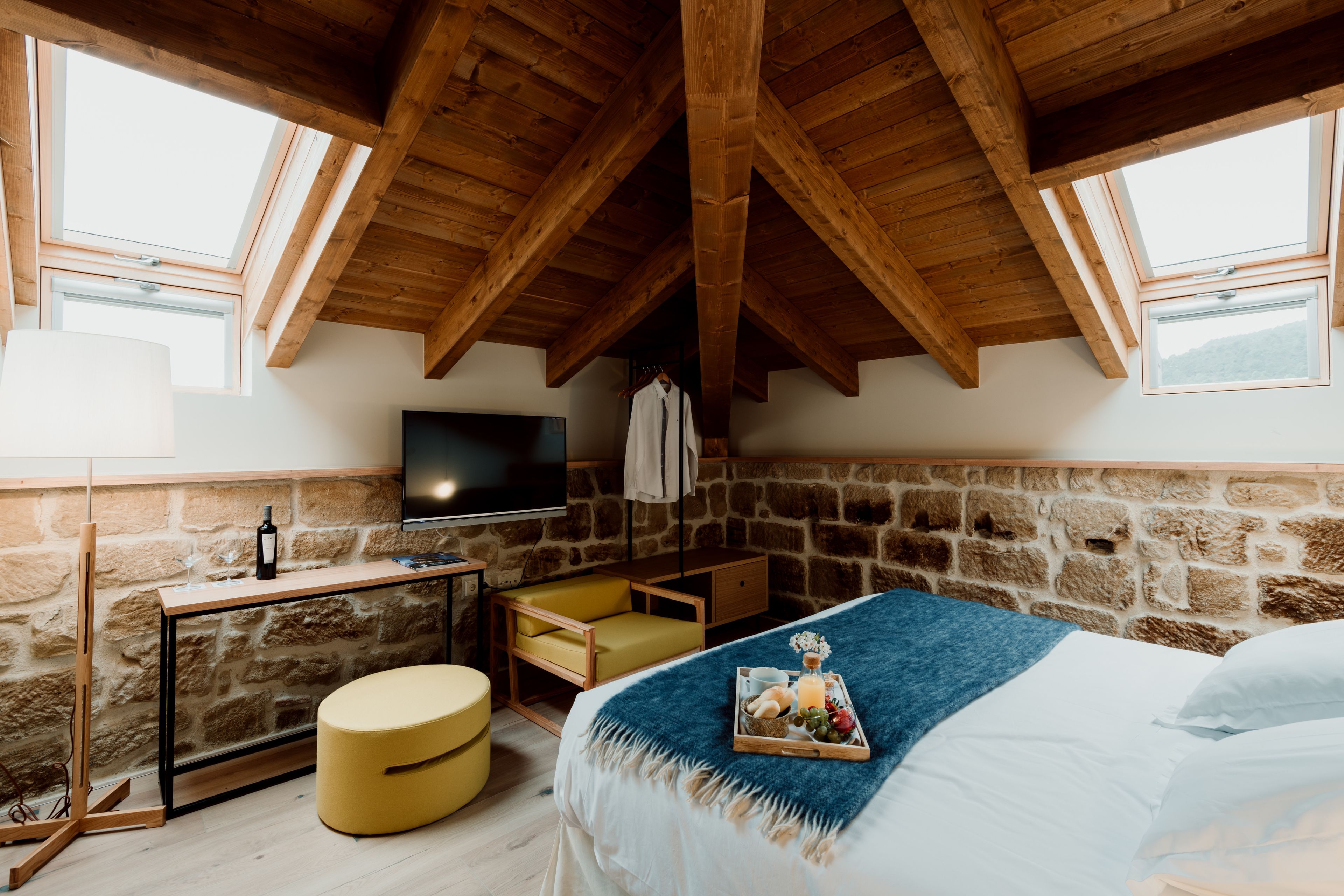 A cozy room with a stone wall, wooden ceiling beams, a bed with a blue blanket, a desk with a wine bottle, and a TV on the wall.