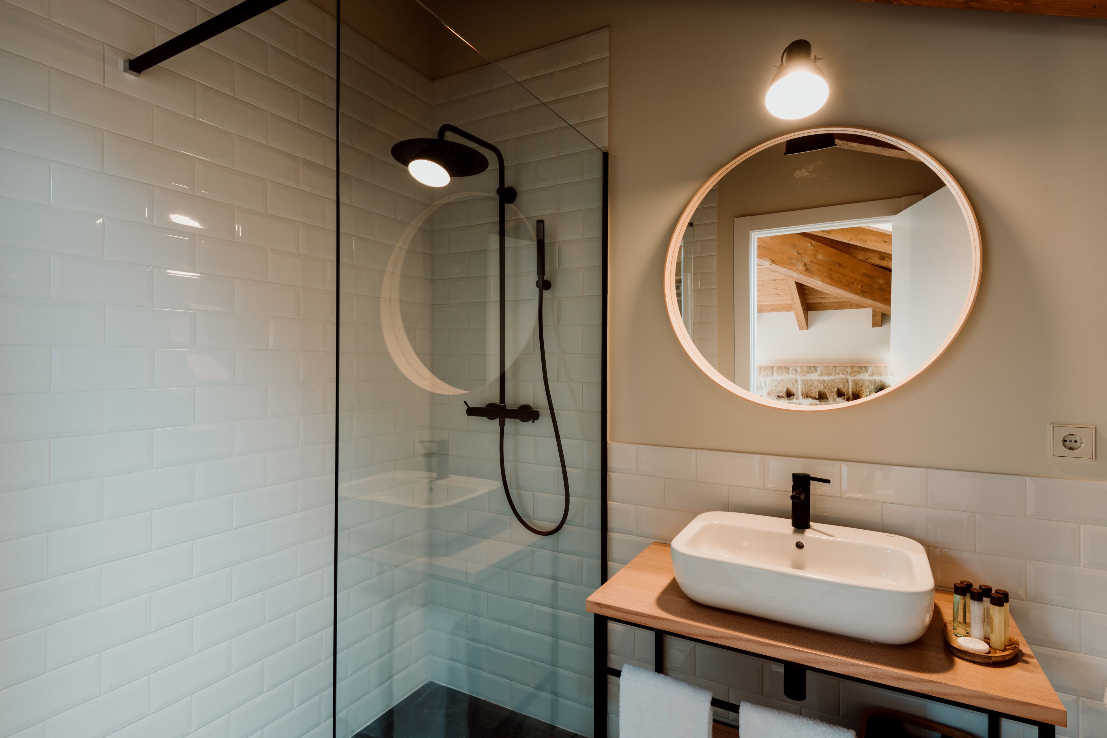 Une salle de bain moderne avec une douche à parois de verre, un miroir rond et un meuble-lavabo en bois avec un évier blanc.