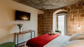 Un dormitorio con pared de piedra, cama con ropa de cama blanca, una manta roja, una mesita de noche con un jarrón de flores y un televisor en la pared.