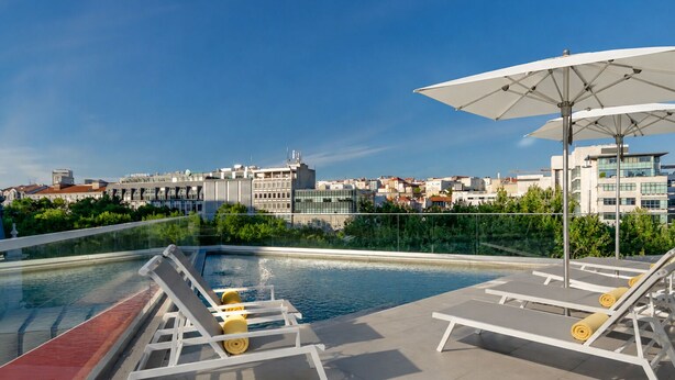 Una piscina en la azotea con sillones y sombrillas, rodeada de vegetación y edificios.
