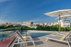 Una piscina en la azotea con sillones y sombrillas, rodeada de vegetación y edificios.