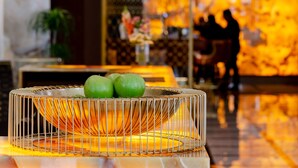 Zona de recepción de un hotel con un bol de frutas metálico sobre una mesa, pared de mármol y un fondo desenfocado con dos personas.