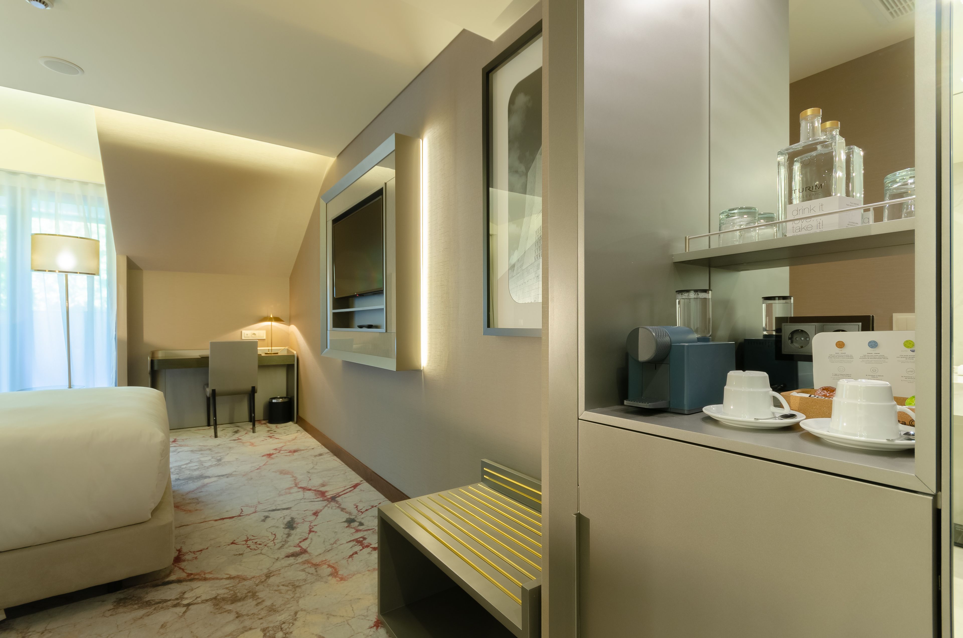 Habitación de hotel con cama, escritorio, silla, banco, espejo, estante con artículos de aseo y cafetera.