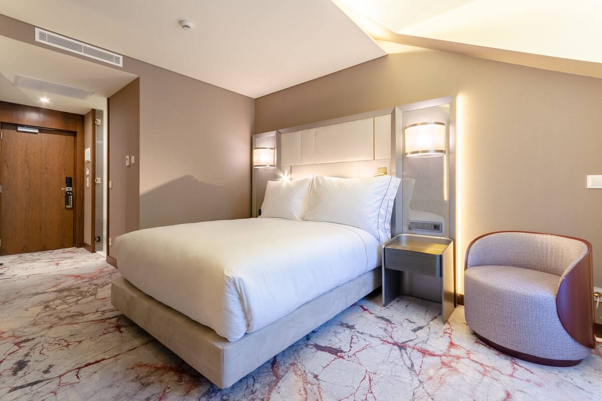 A hotel room with a large bed, a nightstand, a chair, and a marble-patterned floor.
