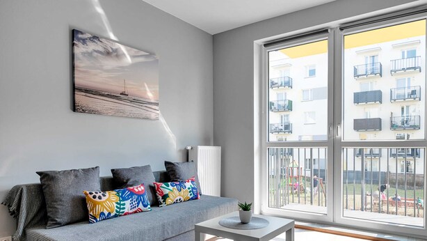 A modern living room with a grey sofa, colorful cushions, a white coffee table, and a large window overlooking a residential area. - Warszawa-Wlochy Brilliant Apartment (Warsaw)