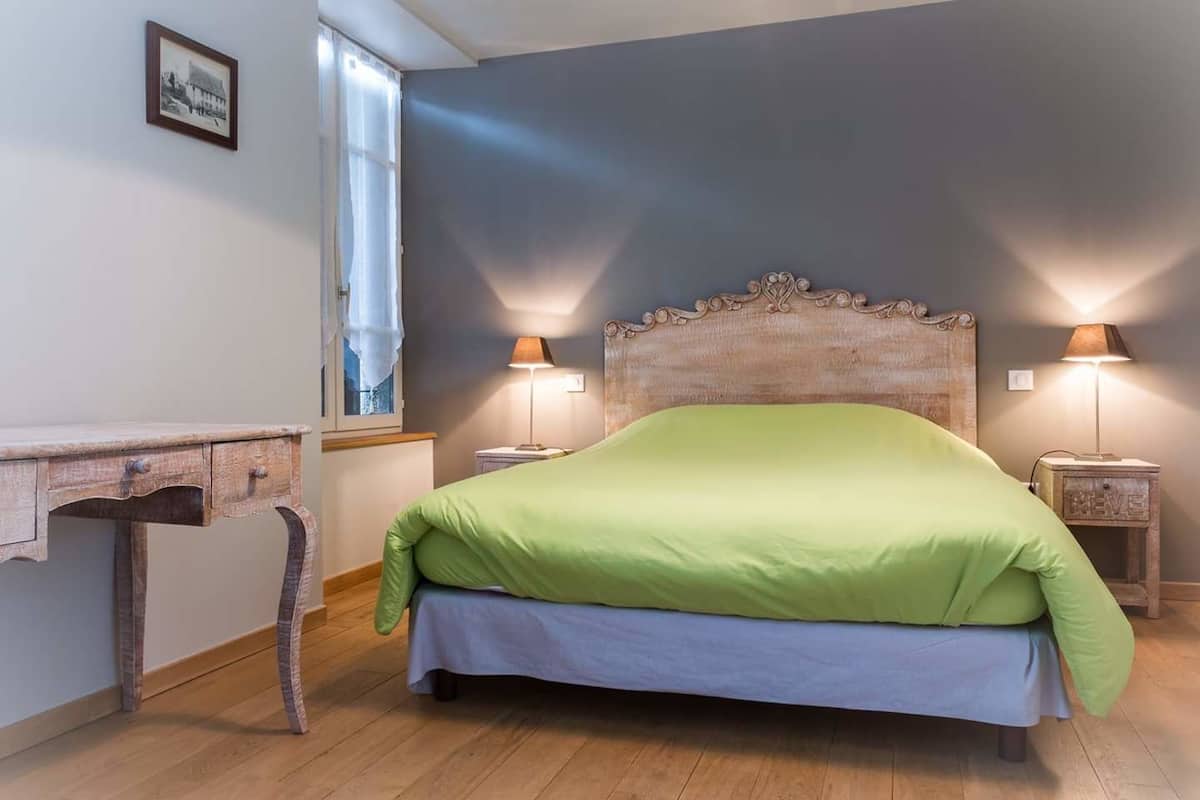 A bedroom with a wooden headboard, a green bedspread, two bedside lamps, a small wooden nightstand, a window with curtains, and a picture on the wall.