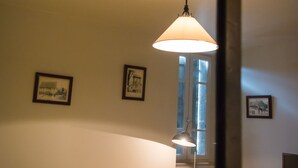 A room with a table, wicker chair, and a lamp. There are framed pictures on the wall and a window with a view. - Auberge de Teissieres (Teissières-lès-Bouliès)