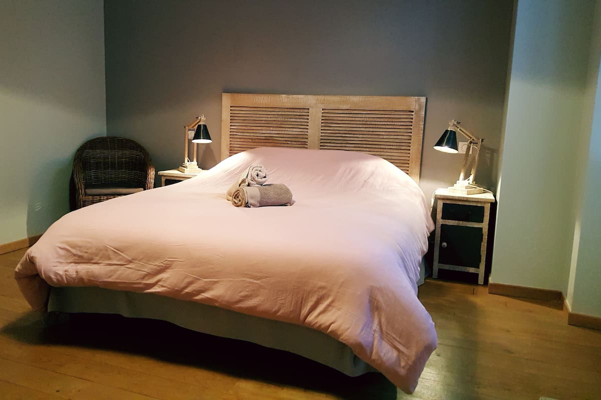 A neatly made bed with a pink bedspread, two bedside lamps, a wicker chair, and a wooden headboard.