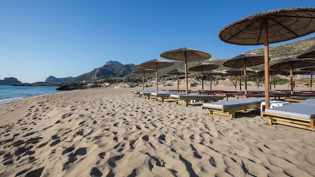 A beach with rows of sunbeds and thatched umbrellas, a clear sky, and mountains in the background. - En Plo Rooms (Kissamos)