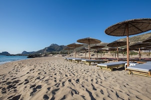 A beach with rows of sunbeds and thatched umbrellas, a clear sky, and mountains in the background.