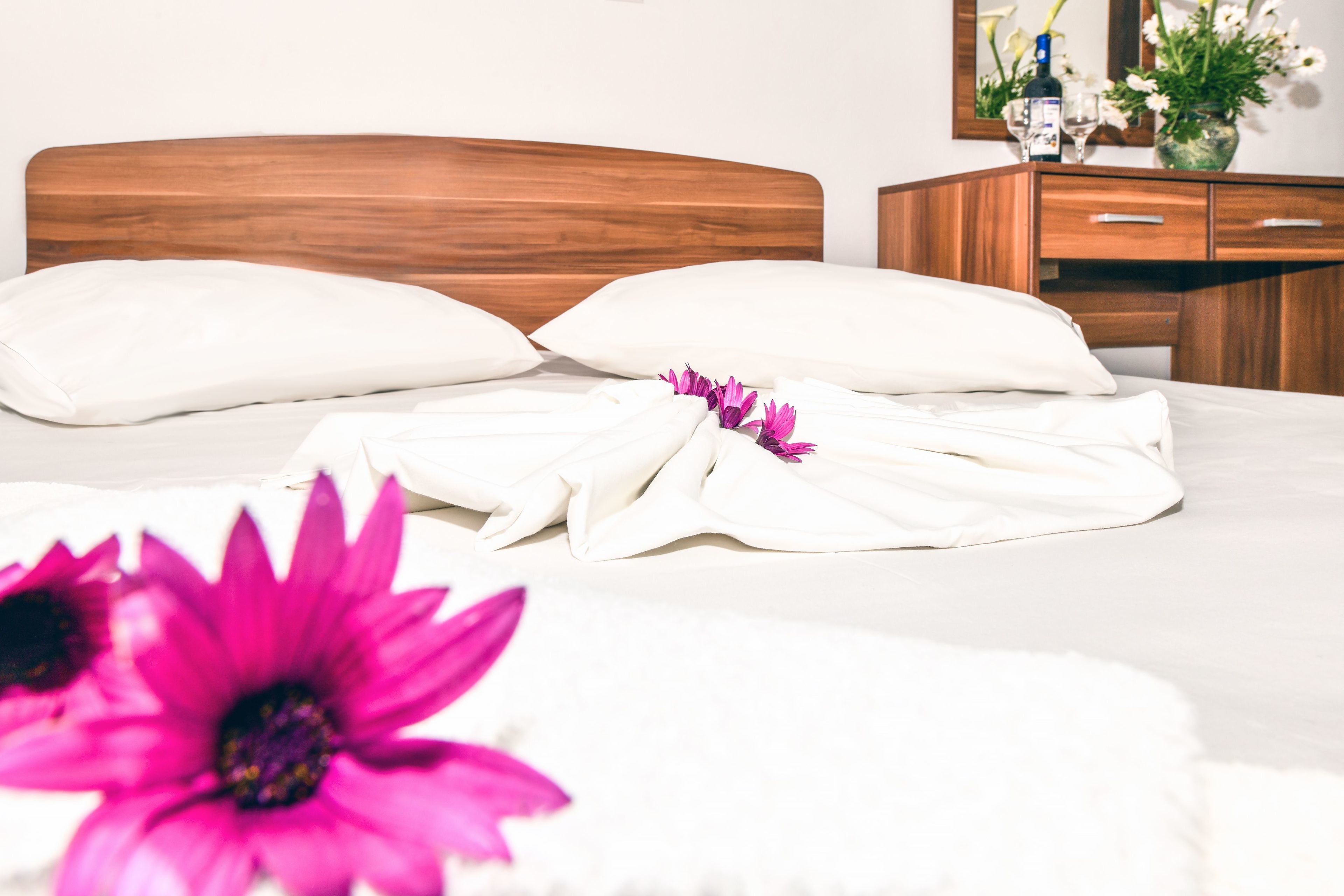 A neatly made bed with white bedding and a wooden headboard, a wooden dresser with drawers, and a vase with flowers on the dresser.