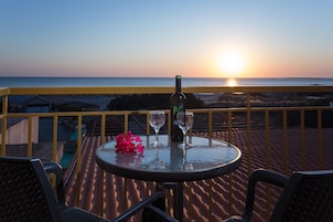 A table with a bottle and two glasses on a balcony overlooking the ocean at sunset.