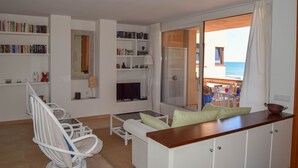 Un salon moderne avec un canapé, une table basse en verre, une bibliothèque et une vue sur la mer, visible à travers une porte-fenêtre coulissante.