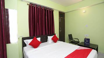 Habitación de hotel con una cama, ropa de cama blanca y roja, un cabecero negro, una ventana con cortinas, una puerta, una mesita con un cartel y una silla.