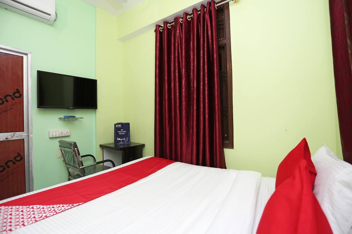 Habitación de hotel con cama, televisor montado en la pared, escritorio con computadora y ventana con cortinas rojas.