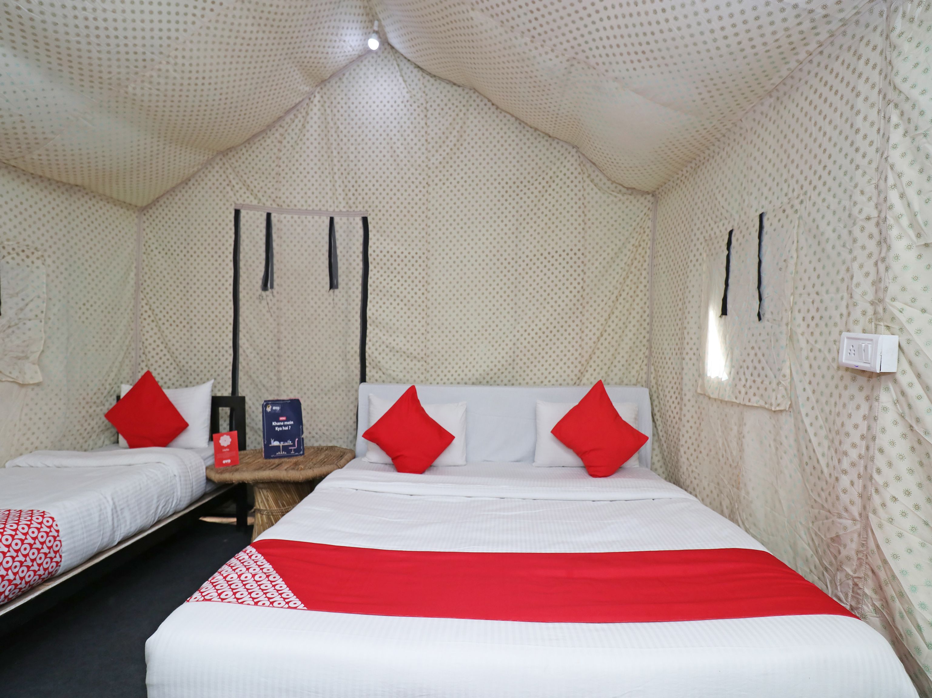 Une chambre avec deux lits, chacun étant équipé de draps blancs et d’oreillers rouges. On y trouve une petite table sur laquelle sont posés une bouteille et une boîte. La chambre a un mur à motifs et un plafond agrémenté d’un luminaire.