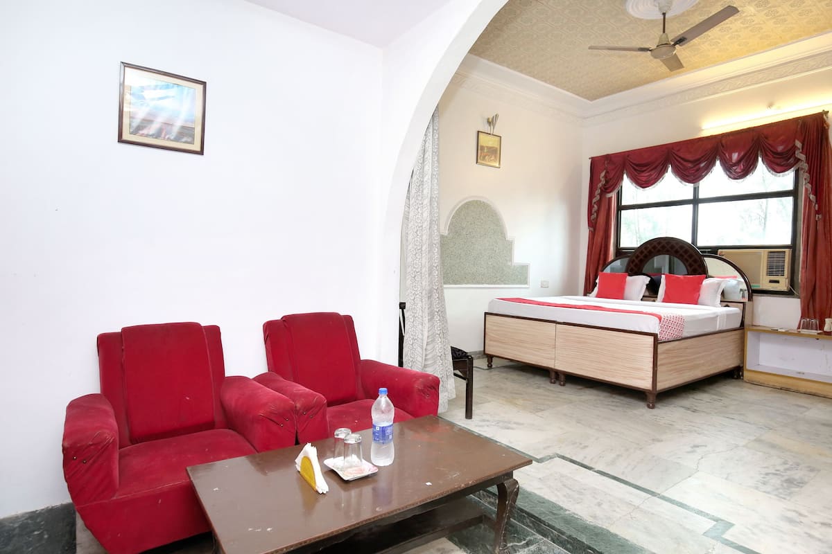 A hotel room with a red sofa, a wooden bed with red and white bedding, a small table with a bottle and glasses, and a ceiling fan.