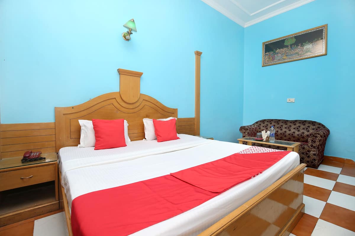 A hotel room with a wooden bed, red and white bedding, a patterned armchair, and a painting on the wall.