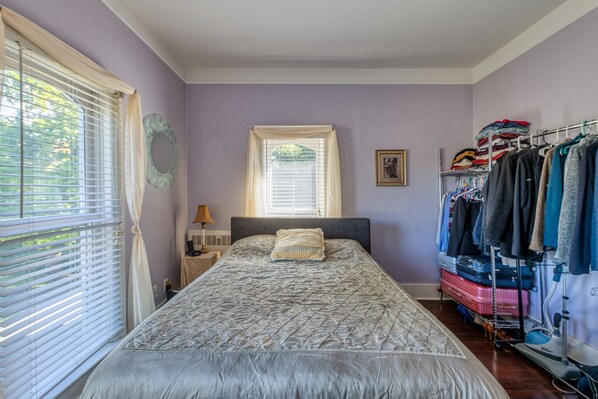 A bedroom with a bed, a window with blinds, a bedside table with a lamp, and a clothing rack with folded clothes. - Monroe Manor Inn (South Haven)