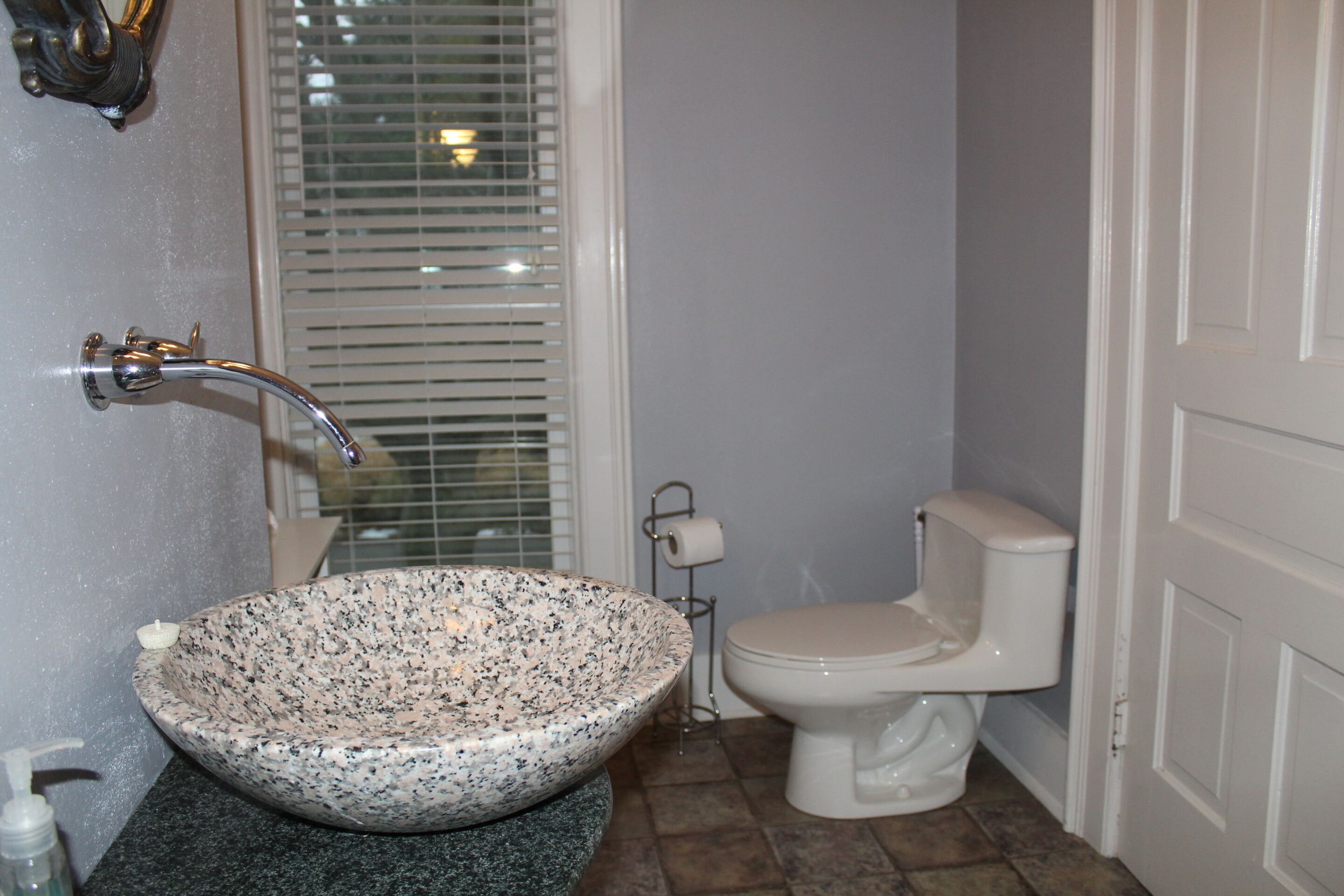 A bathroom with a granite sink, a toilet, and a window with blinds.