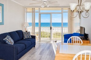 A room with a blue sofa, a dining table with chairs, a ceiling fan, and a view of the ocean through large glass doors. - Grand Strand Resorts II 19 (North Myrtle Beach)