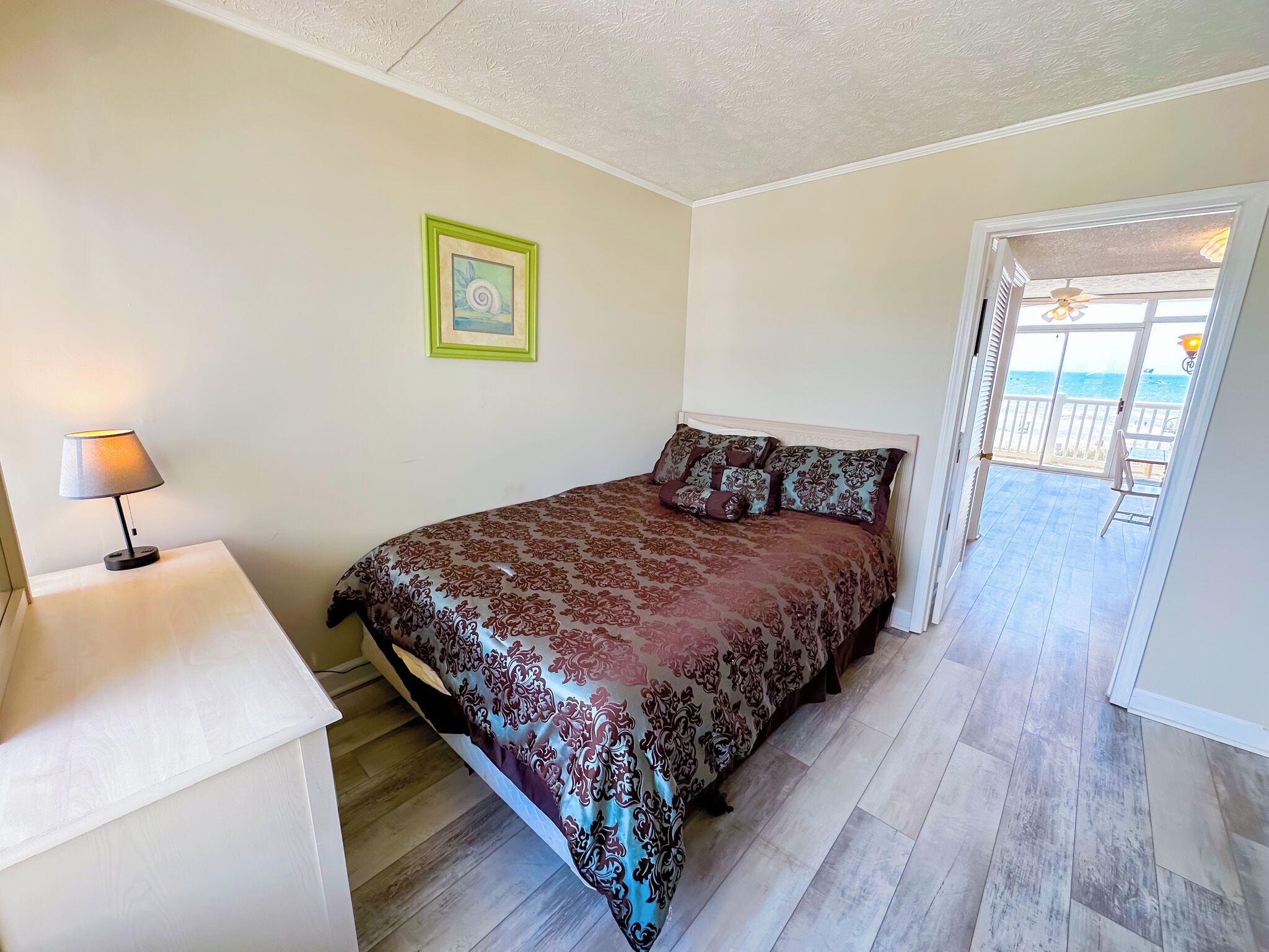 Une chambre à coucher avec un lit, une table de chevet, une lampe, un tableau encadré au mur et une vue sur la mer par une porte ouverte.