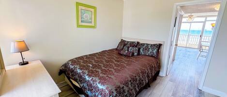 Une chambre à coucher avec un lit, une table de chevet, une lampe, un tableau encadré au mur et une vue sur la mer par une porte ouverte.