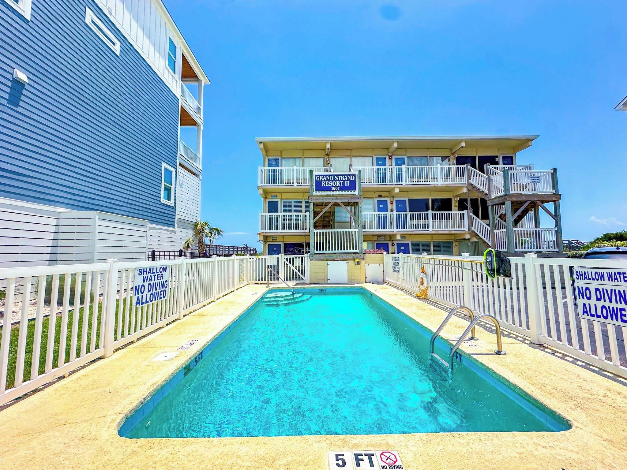 A swimming pool with a 5 FT depth marker, surrounded by a white fence with 'SHALLOW WATER NO DIVING ALLOWED' signs, and a building labeled 'GRAND STRAND RESORT II' in the background.