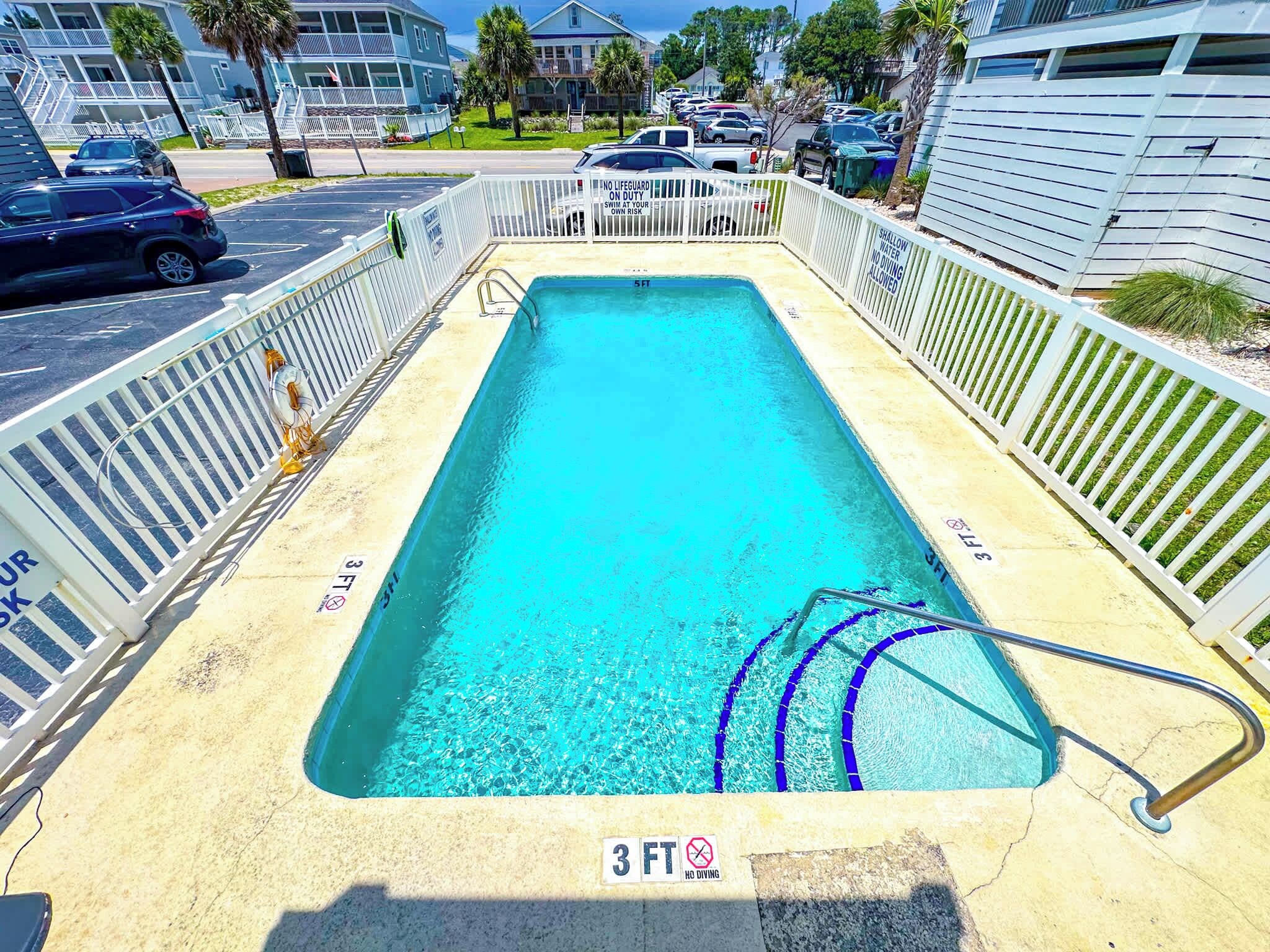 Ein rechteckiger Swimmingpool mit einer Tiefenmarkierung von 3 Fuß am Boden, umgeben von einem weißen Zaun mit einem Warnschild vor flachem Wasser.