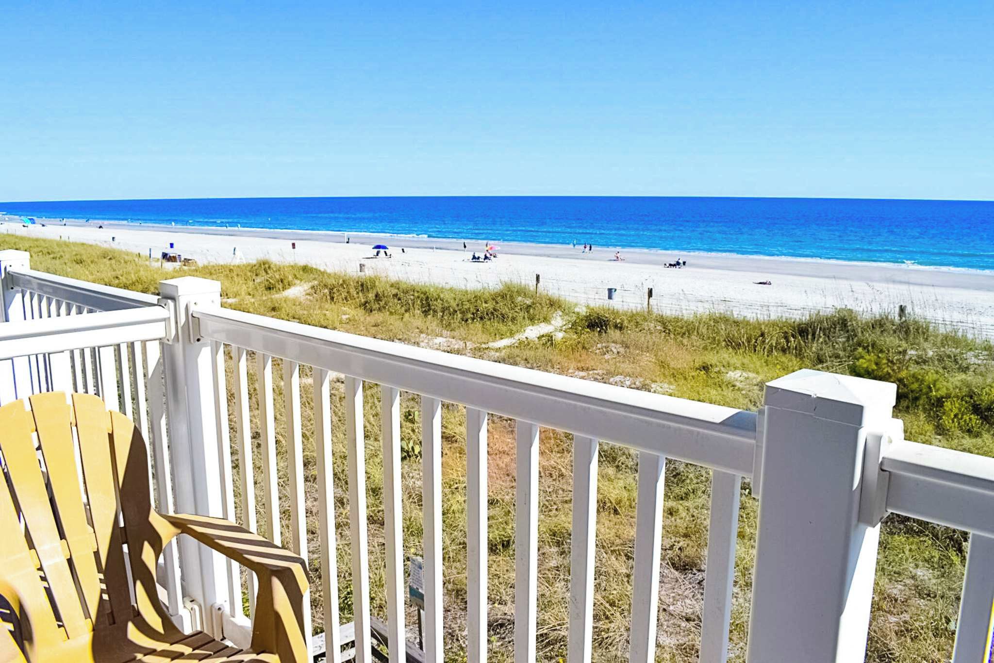 Vista de una playa desde una terraza de madera con barandilla blanca, con vista a una playa arenosa y el océano.