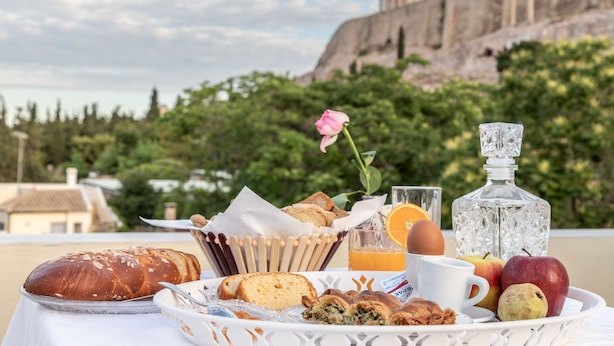A breakfast tray with bread, pastries, fruits, and a bottle on a table with a view of a historical building and greenery. - Kyria House (Athens)