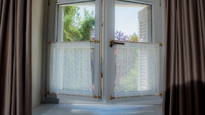 A bay window with white frames and a lace curtain, offering a view of greenery and flowers outside. - Kyria House (Athens)