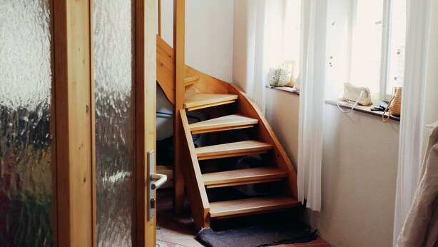 Una scala in legno conduce a un pianerottolo con una finestra e uno scaffale con oggetti.