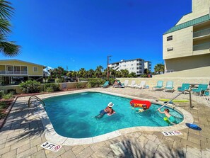 A swimming pool with a 3ft depth marker, lounge chairs, and a building in the background.