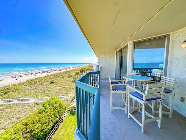 A balcony with a table and chairs, overlooking a beach with umbrellas and people.