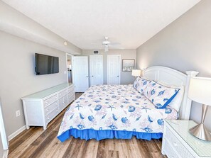 A bedroom with a bed, dresser, ceiling fan, and a TV mounted on the wall.