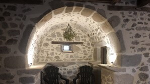 A stone-walled room with two black plastic chairs, a small shelf with a plant, and a window.