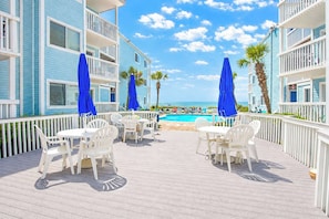A poolside area with white chairs and blue umbrellas, a wooden deck, and a view of the ocean.
