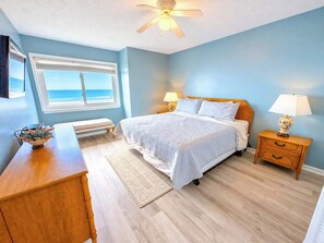 Une chambre à coucher avec un lit, des tables de chevet, un bureau, une vue sur l’océan et un ventilateur de plafond.