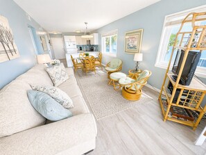 A living room with a sofa, chairs, a TV stand, and a kitchen in the background.
