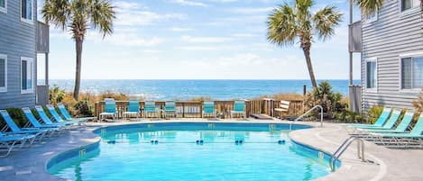 A swimming pool with lounge chairs and palm trees, surrounded by buildings with a view of the ocean.