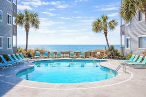 A swimming pool with lounge chairs and palm trees, surrounded by buildings with a view of the ocean.