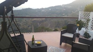 Un balcon avec un fauteuil suspendu, une table avec une bouteille et un saladier, et une vue sur un paysage vallonné.