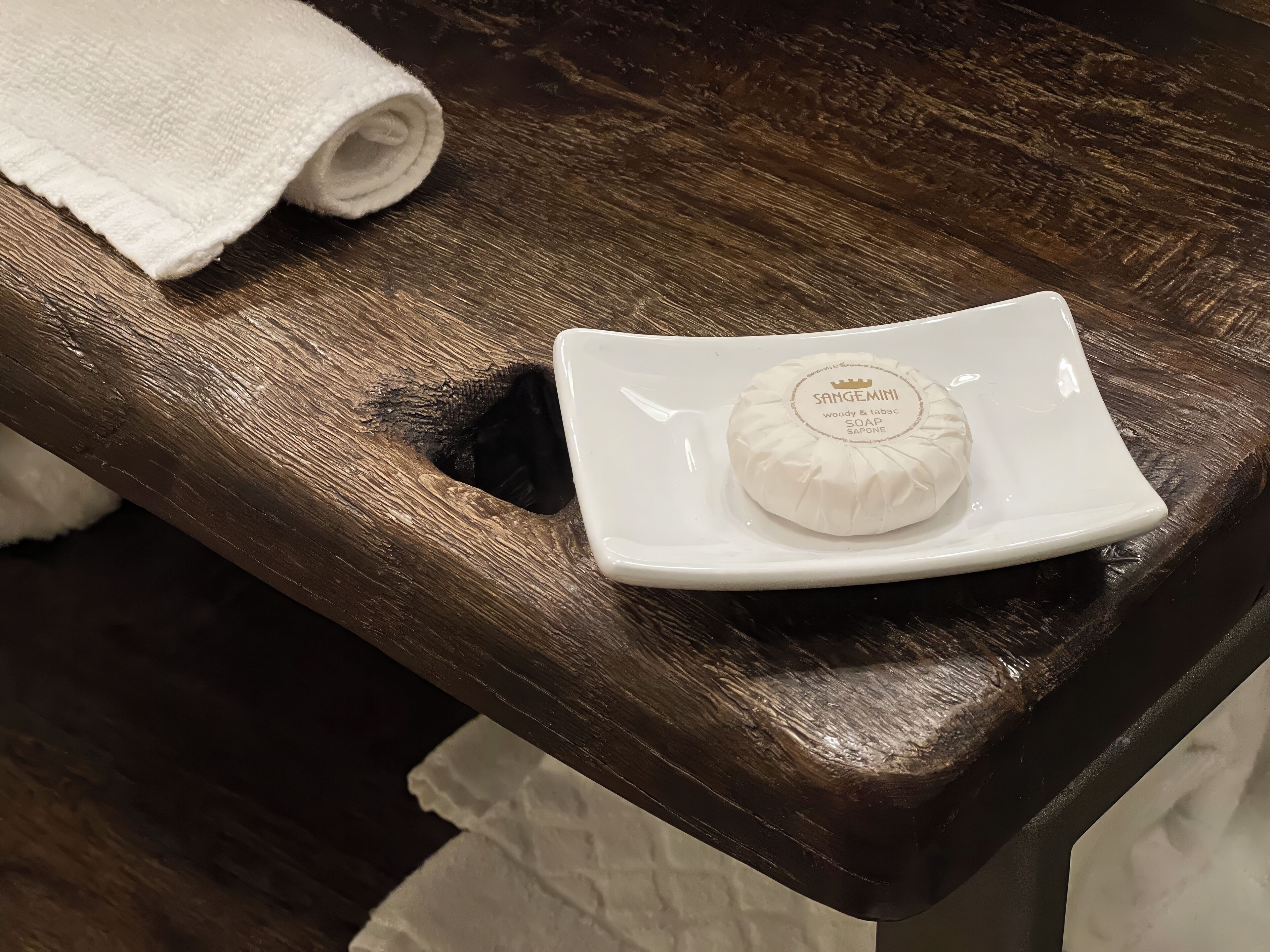 Una pastilla de jabón blanca con el nombre 'SANGEMINI' sobre un plato cuadrado blanco sobre una superficie de madera.