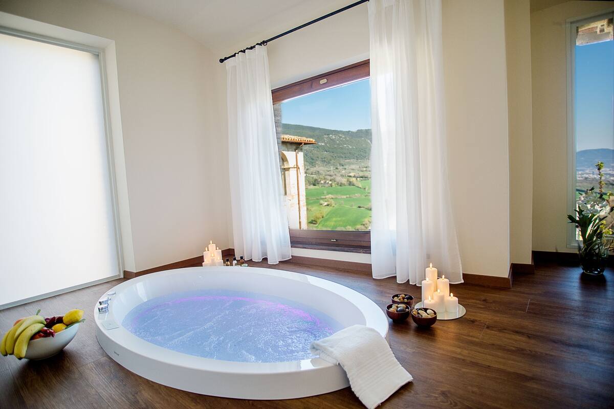 Une salle de spa avec un jacuzzi, un panier de fruits, des bougies et une vue sur un pont et des collines.