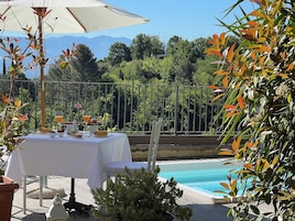 Zona de comedor junto a la piscina con una mesa para dos, un vaso de jugo de naranja y una planta pequeña.