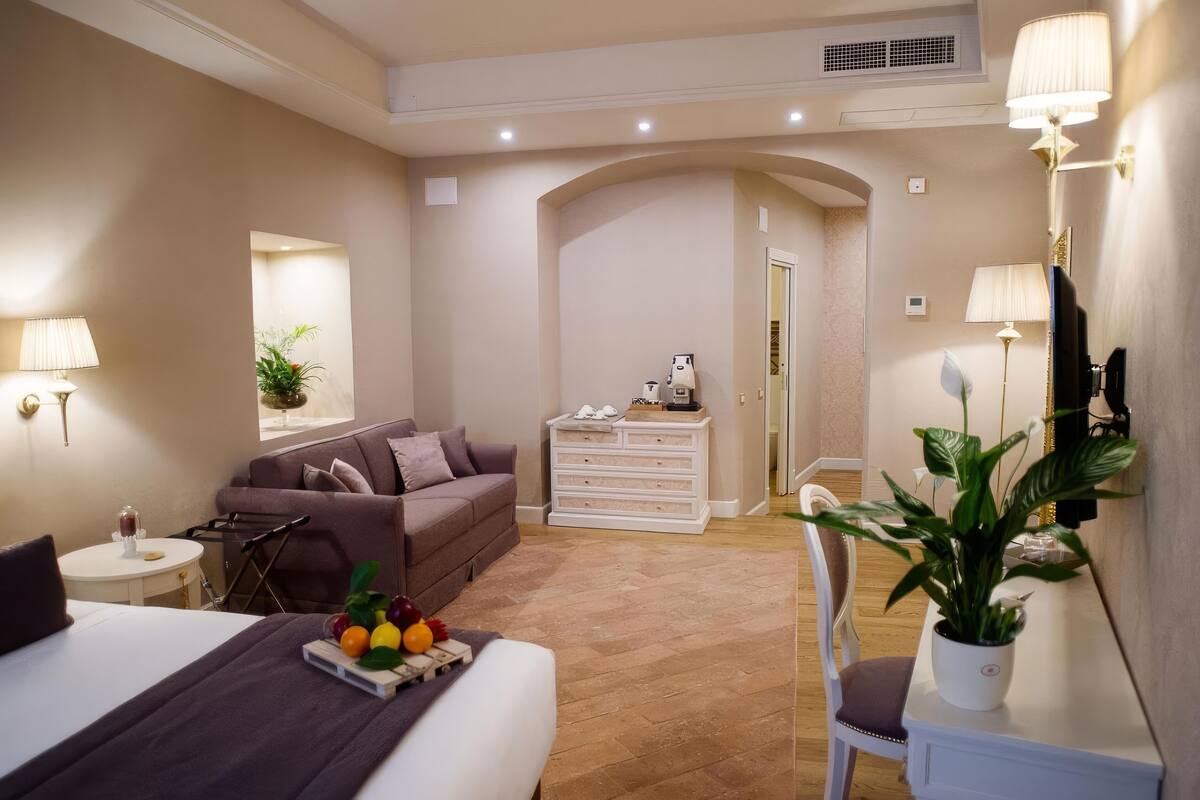 Une chambre d’hôtel moderne, équipée d’un lit, d’un canapé, d’une petite table avec des fruits, d’une commode et d’une plante en pot.
