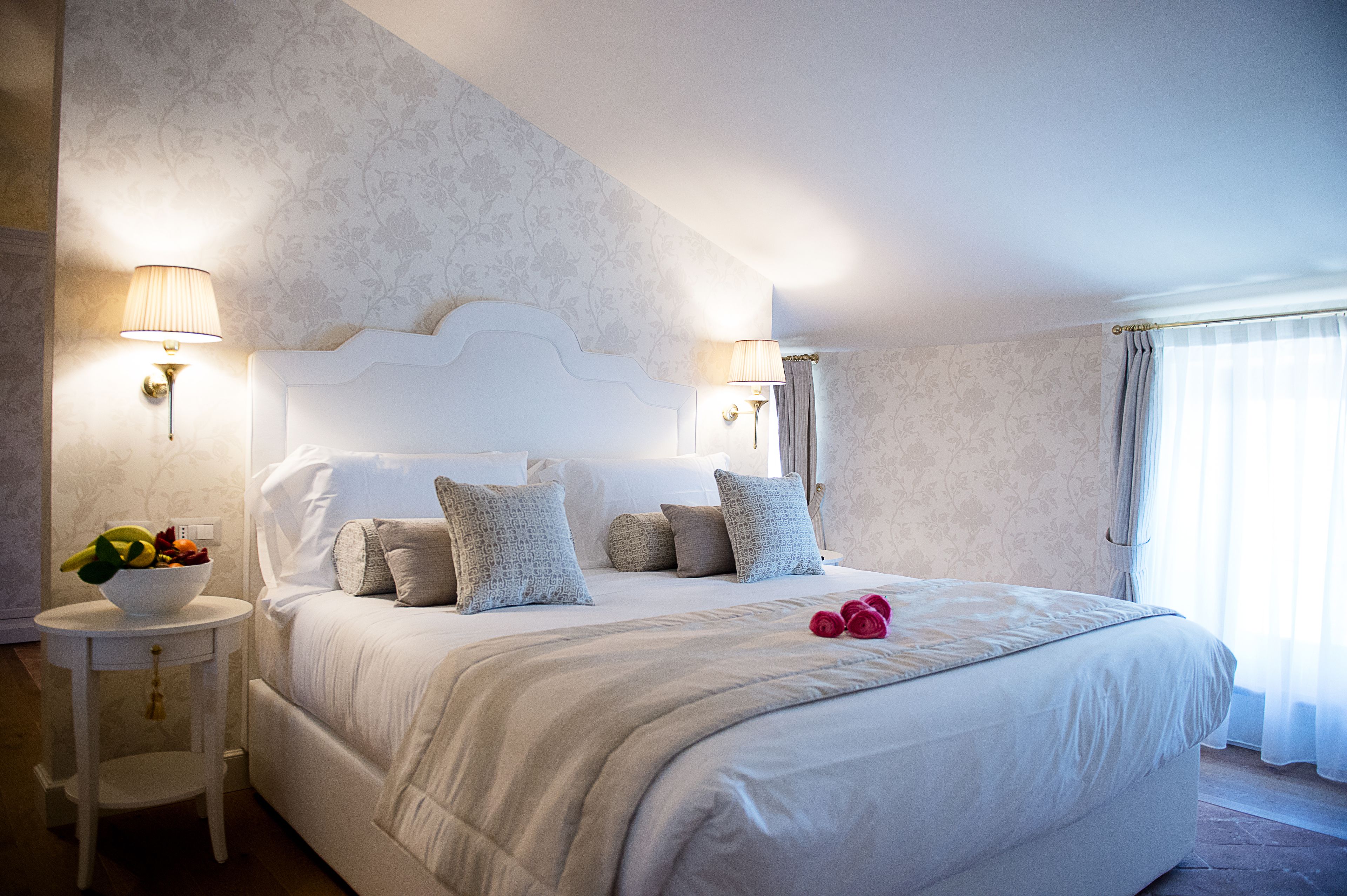 A neatly made bed with a white headboard, two bedside lamps, and a bowl of fruit on a small table beside the bed.
