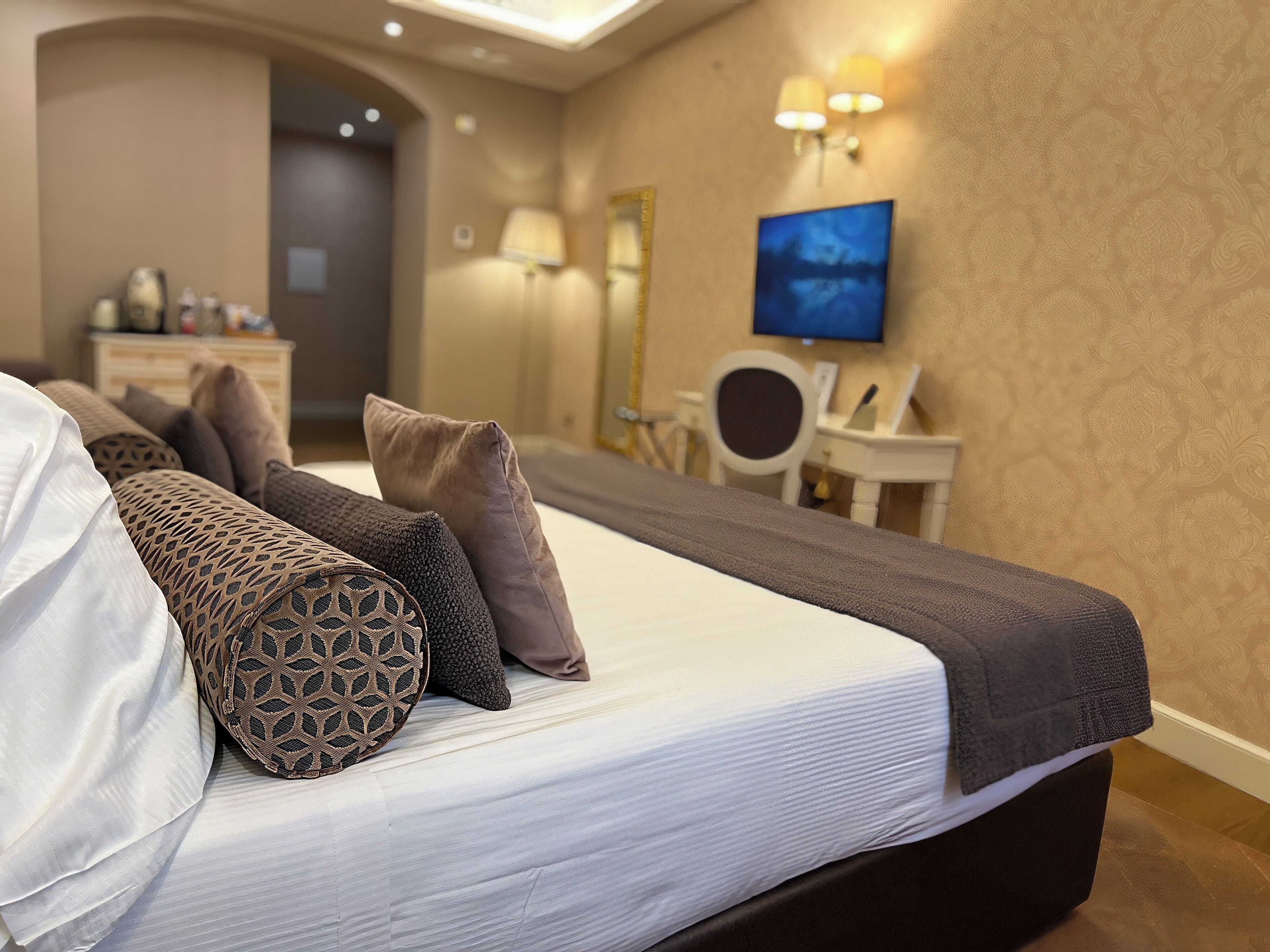 Ein ordentlich bezogenes Bett mit gemusterten Kissen, ein Nachttisch mit Lampe und ein Schreibtisch mit Stuhl in einem Hotelzimmer.