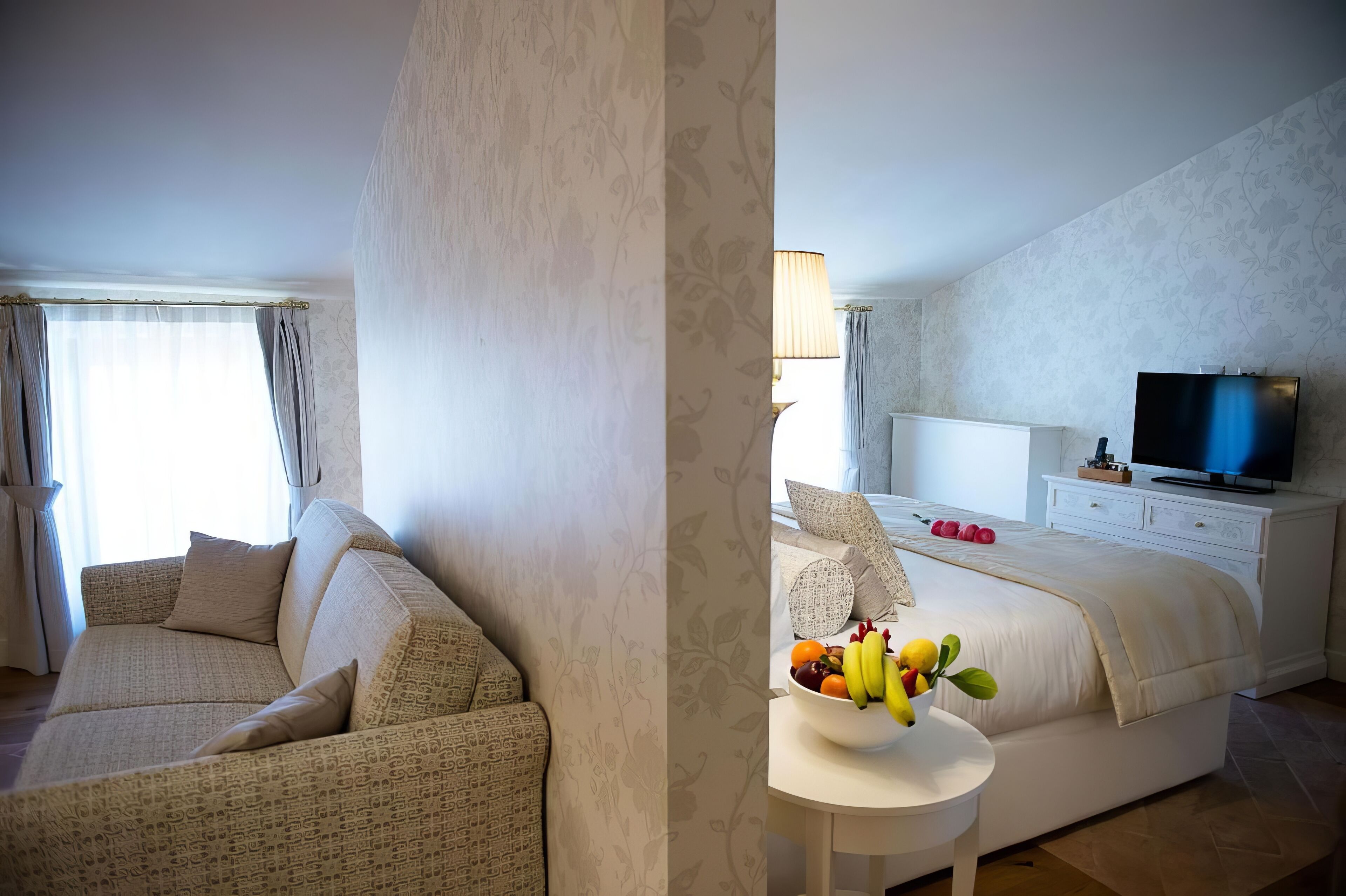 A hotel room with a sofa, a bed with a TV, and a small table with fruit.